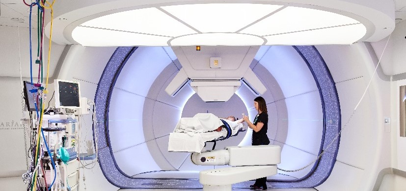 People modeling roles of a technician and patient preparing for proton therapy treatment in a bright white room with a box-like proton delivery device that can rotate in a full circle around the patient.