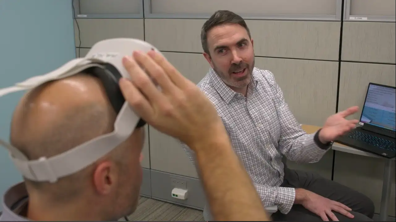 Two experts at Cincinnati Children's, one removing a virtual reality headset, the other sitting at a laptop computer, discuss a virtual reality training program that can help clinicians speak more effectively with families