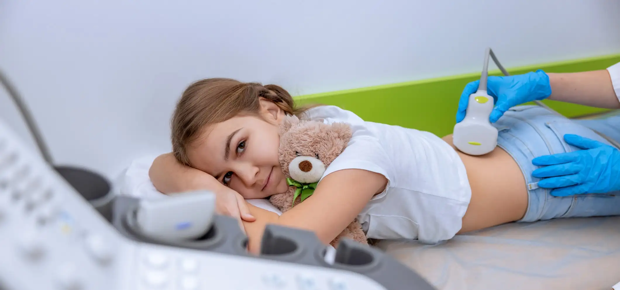 Child wearing a white shirt and blue shorts lies on an exam table holding a brown teddy bear while a healthcare provider wearing blue gloves performs an abdominal ultrasound with a handheld imaging device. An ultrasound machine is visible in the foreground, with white and green walls in the background.