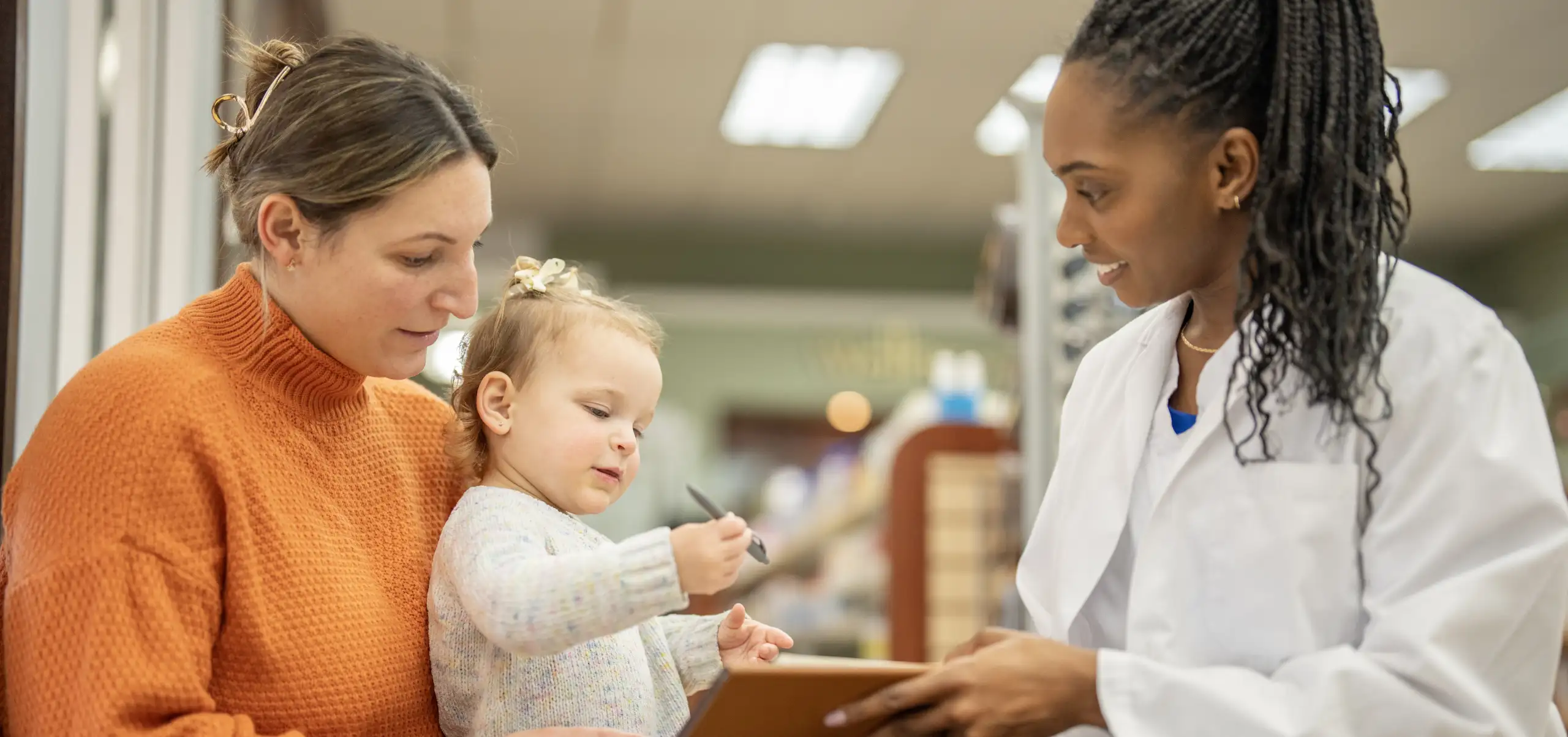 A healthcare provider reviews treatment options with a caregiver and young child during a pediatric urology clinic visit. The child holds a pen while looking toward a clipboard or tablet presented by the provider. The image illustrates a conversation about care planning and medical management in a bright outpatient healthcare setting.